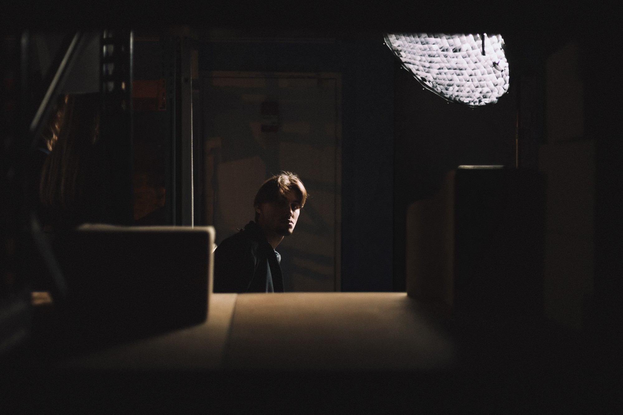 Langa, à moitié dans l’ombre, regarde l’objectif derrière une table, sous la lumière d’une boîte à lumière de studio